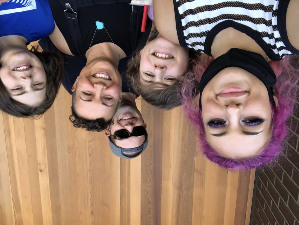 Wood wall background. Older teen with pink hair, long lashes and purple eyeshadow, younger teen with shaggy brown hair, middle-age man with backwards hat and sunglasses, middle-age woman with short curly hair, and young teen with shaggy brown hair - all smiling. Tag: fear self-care strategies