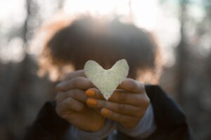 woman holding a yellow, heart shaped leaf with orange nail polish in the forefront of the picture and woman's head and trees are blurred in the background Tag: 2020 reflection gratitude grief
