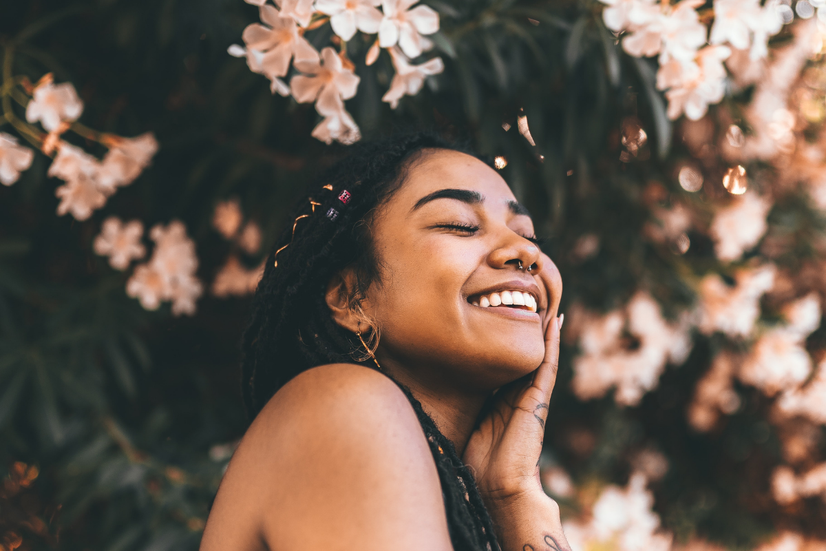 brown skinned woman standing near a pink flower tree smiling with her eyes closed, Tag: what others think of you