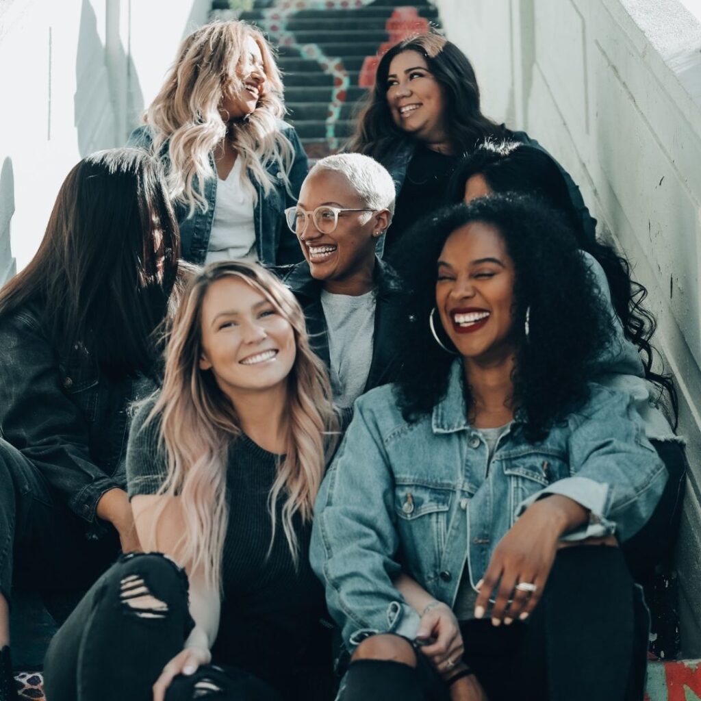 Group of women sitting close together on stairs, smiling at each other. Front row: white woman with long blonde hair wearing a black short sleeve shirt sitting beside woman with long black curly hair, brown skin, wearing a blue jean jacket. Second row: woman with long black hair, brown skin, wearing long sleeve black shirt looking at woman with platinum blonde hair, brown skin, glasses and white shirt. Third row: white woman with long blonde hair wearing white shirt and blue jean jacket smiling and looking at woman with long black hair, brown skin, wearing black shirt. Image Credit: Joel Muniz; tag: may 2022 self-care